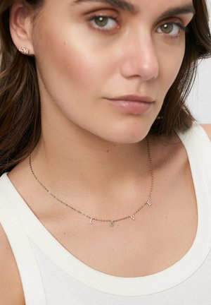 Collana in oro rosa con le lettere "LOVE" impreziosite da pietre trasparenti; il modello indossa una canotta bianca. Gli orecchini riportano la parola "LOVE".