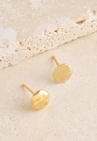 Goldene Ohrstecker mit glatter, unregelmäßiger runder Form und glänzendem Finish. Auf einer strukturierten, hellen Oberfläche platziert.