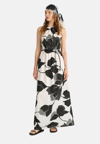 Abito maxi bianco con grandi motivi di foglie nere e grigie. Presenta un colletto alto, vita elastica e un foulard abbinato. Sandali piatti.