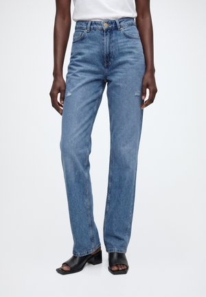 Underkrop iført blå højtaljede jeans med let slid, kombineret med sorte blokhælede sko med åben tå og en hvid top.