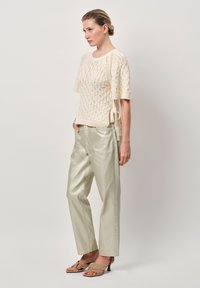 Gehaakte crèmekleurige top met korte mouwen en een opengewerkte patronen, gecombineerd met lichtmetallisch groene broek. Sandalen hebben franjes.