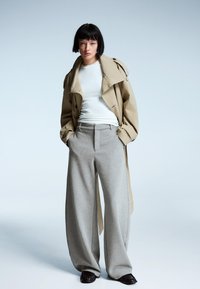 Beige trenchcoat over een witte shirt, gecombineerd met hoogwaardig, wijde grijze broek en zwarte platte schoenen. Glad stoffeerwerk.
