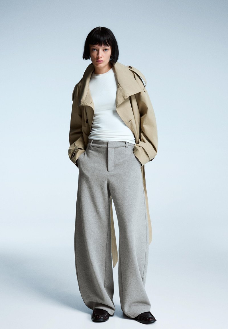 Beige trenchcoat over een witte shirt, gecombineerd met hoogwaardig, wijde grijze broek en zwarte platte schoenen. Glad stoffeerwerk.