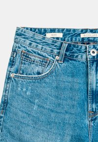 Jeans de denim en azul claro con una textura desvanecida, que presentan un bolsillo frontal, trabillas para el cinturón y herrajes de metal. Etiquetas visibles en la cinturilla.