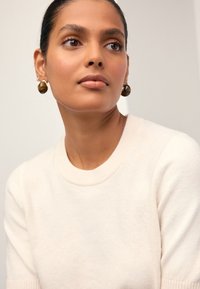 Pull crème à col rond et manches courtes, associé à de grandes boucles d'oreilles rondes brunes. Texture lisse, design minimaliste.