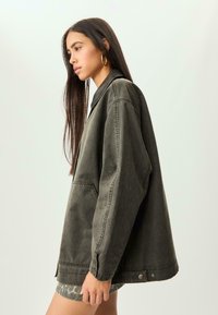 Giacca di denim oversize verde oliva con colletto nero, tasca frontale e dettaglio con bottoni a pressione sull'orlo, mostrata di profilo.