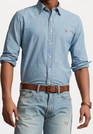 Mand iført lyseblå skjorte med knapper og opsmøgede ærmer, lyse vaskede jeans, brun læderbælte og brun ur på venstre håndled.