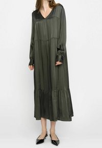Robe à manches longues, décolleté en V de couleur vert foncé, présentant une texture plissée, un ourlet à étages et des manches à poignet. Portée avec des chaussures noires à bout pointu.