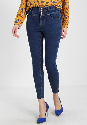 Jean en denim foncé ajusté avec taille haute, fermeture à cinq boutons et ourlet effiloché. Porté avec un haut à fleurs et des talons bleus.