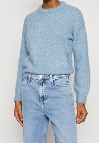 Hellblauer, kuscheliger Pullover mit Rundhalsausschnitt und langen Ärmeln, der eine glitzernde Textur aufweist. Kombiniert mit hellblauen Jeans.