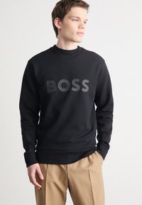 Zwarte sweatshirt met ronde hals, voorzien van een matte "BOSS" logo op de voorkant. Gladde textuur, in combinatie met beige broek.