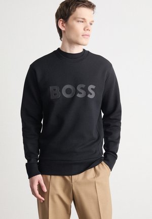Sweatshirt noir à col rond, arborant un logo "BOSS" mat sur le devant. Texture lisse, accompagné de pantalons beiges.