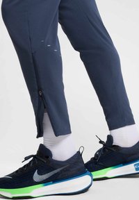 Navy athletic pants with zippered ankle openings; smooth fabric texture. Worn with black and blue Nike shoes featuring green accents.