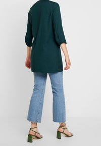 Person iført en mørkegrøn langærmet tunika, lyseblå cropped jeans med frynsede kanter, og grønne strappy block-heeled sandaler, stående.