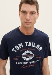 Navy blue cotton t-shirt featuring white and red logo print with "TOM TAILOR," crew neckline, short sleeves, and soft texture.