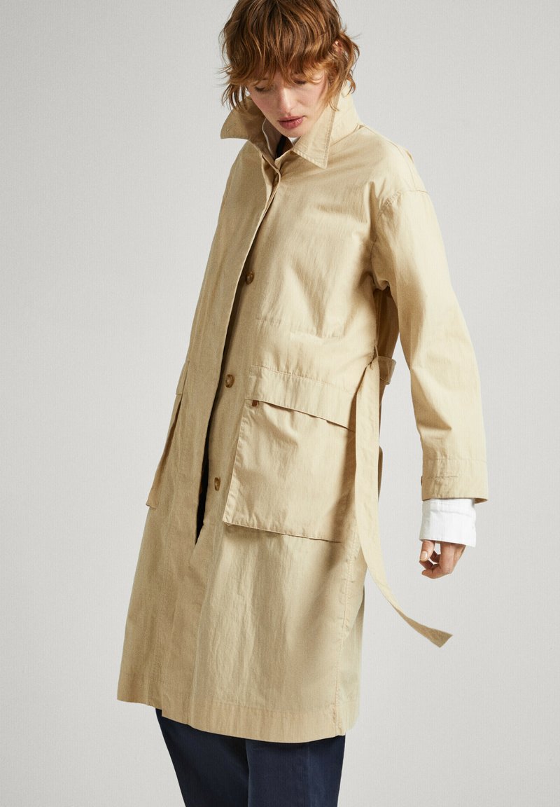 Trench-coat léger beige avec un col, fermeture à boutons, poches latérales et ceinture, en tissu texturé.