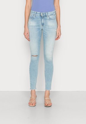 Femme portant un jean skinny bleu clair déchiré et des sandales beiges à brides, debout devant un fond blanc uni.