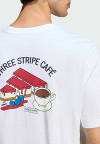 Biela tričko má na zadnej strane farebnú grafiku s dvoma plátkami koláča, šálkou kávy a textom "THREE STRIPE CAFÉ" nad ním.