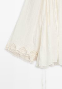 Tessuto bianco fluido con bordo in pizzo smerlato che presenta ritagli geometrici lungo il bordo, appeso contro uno sfondo semplice.