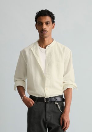 Mann mit lockigem Haar, der ein weißes Hemd mit Knöpfen und schwarze Hose trägt, steht mit einer Hand in der Tasche vor einem einfarbigen Hintergrund.