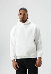 Witte capuchontrui gemaakt van zacht materiaal, met een voorzak en geribbelde boorden, gecombineerd met zwarte jeans.