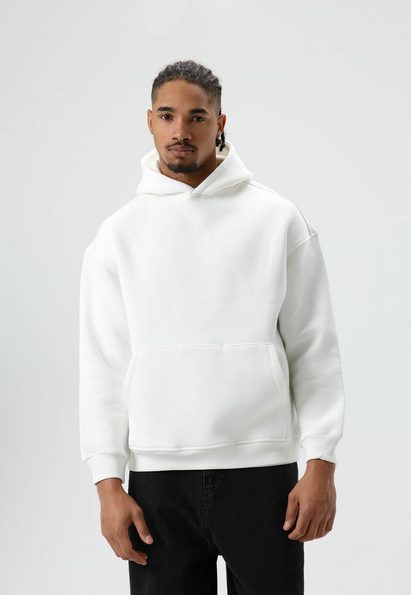 Witte capuchontrui gemaakt van zacht materiaal, met een voorzak en geribbelde boorden, gecombineerd met zwarte jeans.