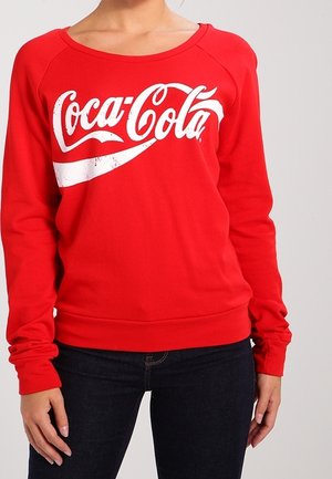 Person trägt einen roten Pullover mit langen Ärmeln und einem weißen Coca-Cola-Logo auf der Brust, kombiniert mit dunkelblauen Jeans.