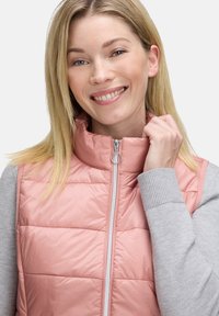 Jasnoróżowy puchowy kamizelka z wysokim kołnierzem i zamkiem z przodu, w zestawie z szarym, długim topem z podwiniętymi mankietami.