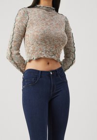 Suéter de punto con un diseño corto en tonos terrosos y bordes en forma de concha, combinado con jeans ajustados de color azul oscuro que tienen bolsillos frontales.