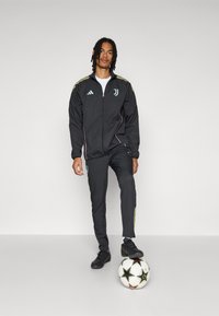 Ensemble de survêtement sportif noir avec veste zippée et pantalons longs, ornés de rayures jaunes et d'un logo bleu clair, associé à des baskets noires et à un ballon de football.