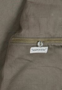 Alyvuogių žalios spalvos audinys su lygiu paviršiumi, su siuvinėta etikete, kurioje parašyta „Iki mirties mus skiria“, pritvirtinta suapvalintu mygtuku.