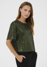 Olijfgroene top met pailletten, korte mouwen, ronde halslijn en losse pasvorm, gedragen met zwarte broek. Gladde textuur met glanzende accenten.