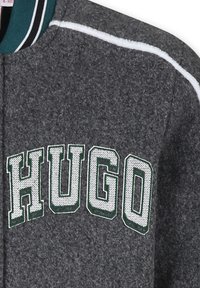 Grijze bomberjack van vilt met een groen en wit geborduurd "HUGO" logo op de borst, voorzien van gestreepte kraagaccenten.