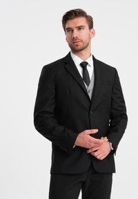 Musta puku takki, jossa on viilletty kaulus, yhdistettynä valkoiseen paitaan ja mustaan solmuun. Alla harmaa liivi ja sileä kankaan tekstuuri.