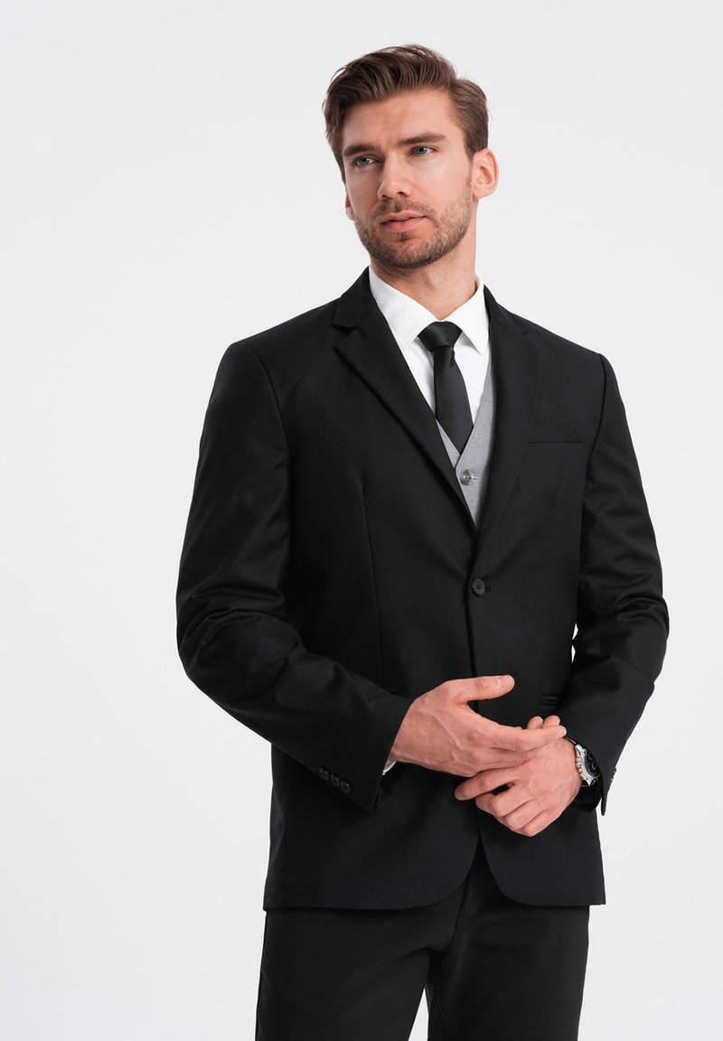 Musta puku takki, jossa on viilletty kaulus, yhdistettynä valkoiseen paitaan ja mustaan solmuun. Alla harmaa liivi ja sileä kankaan tekstuuri.