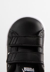 Avant de chaussure de sport Puma noir avec semelle blanche et deux bandes Velcro noires sur fond uni clair.