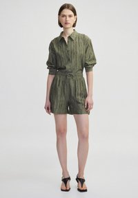 Rompers a maniche corte di colore verde oliva con colletto a bottoni, tessuto testurizzato, vita elasticizzata e tasche laterali. Sandali neri completano il look.