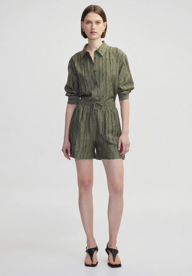 Rompers a maniche corte di colore verde oliva con colletto a bottoni, tessuto testurizzato, vita elasticizzata e tasche laterali. Sandali neri completano il look.