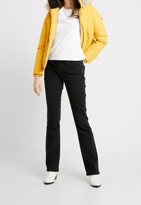 Una mujer se presenta con confianza, vistiendo una chaqueta acolchada amarilla, una camiseta blanca, jeans negros y botas blancas hasta el tobillo, frente a un fondo liso.