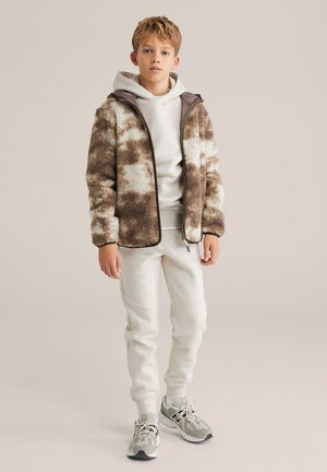 Jachetă din fleece cu un model maro și alb, purtată peste un hanorac alb și pantaloni de trening gri deschis, asortați cu adidași gri. Textură netedă.