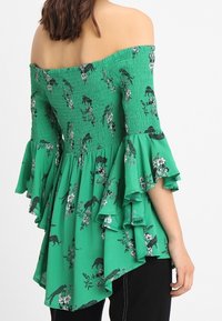 Top verde con spalle scoperte, corpino arricciato, maniche a campana e motivi floreali che presentano leopardi neri e fiori bianchi.