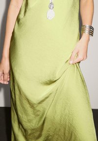 Grünes, fließendes Kleid mit glatter Textur. Trägt eine silberne Halskette und ein silbernes Armband am Handgelenk. Die Hand hält den Stoff des Kleides.