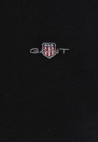 Μαύρο ύφασμα με μικρό κεντημένο λογότυπο GANT που απεικονίζει μια ασπίδα με λευκά αστέρια σε μπλε φόντο και κόκκινες και λευκές ρίγες.