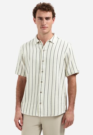 Mann trägt ein kurzärmeliges, weißes Hemd mit schwarzen vertikalen Streifen und beigen Hosen vor einem schlichten Hintergrund.