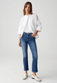 Camicia bianca con bottoni, jeans blu a gamba dritta e mocassini bianchi. La camicia presenta un design senza colletto e maniche arrotolate.