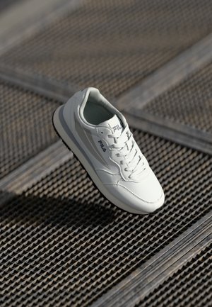 White leather sneaker with grey accents, round toe, padded collar, lace-up design, and textured black rubber sole. Brand logo on side.