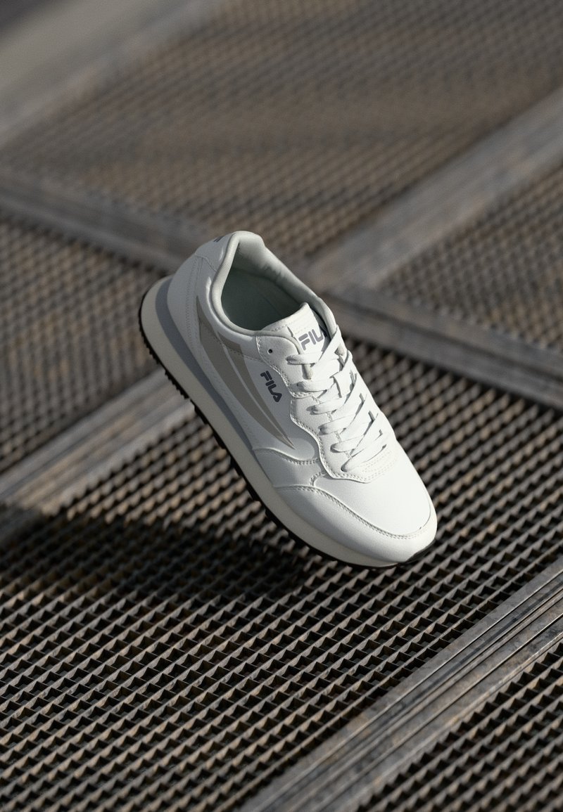 White leather sneaker with grey accents, round toe, padded collar, lace-up design, and textured black rubber sole. Brand logo on side.