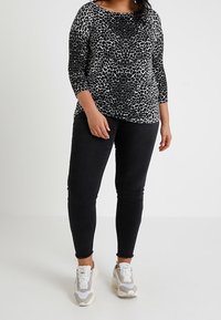 Žena v černobíle leopardím potisku, černých skinny džínách a bílých teniskách na jednobarevném světlém pozadí.