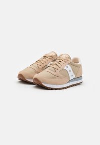 Beige treningssko med semsket skinn og nylonpaneler, hvite detaljer, strukturert snøring og såle av brunt gummi. Side logo detalj synlig.