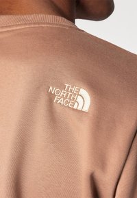 Sudadera marrón usada por una persona, mostrando el logo bordado en blanco de The North Face en la parte superior de la espalda cerca del hombro.